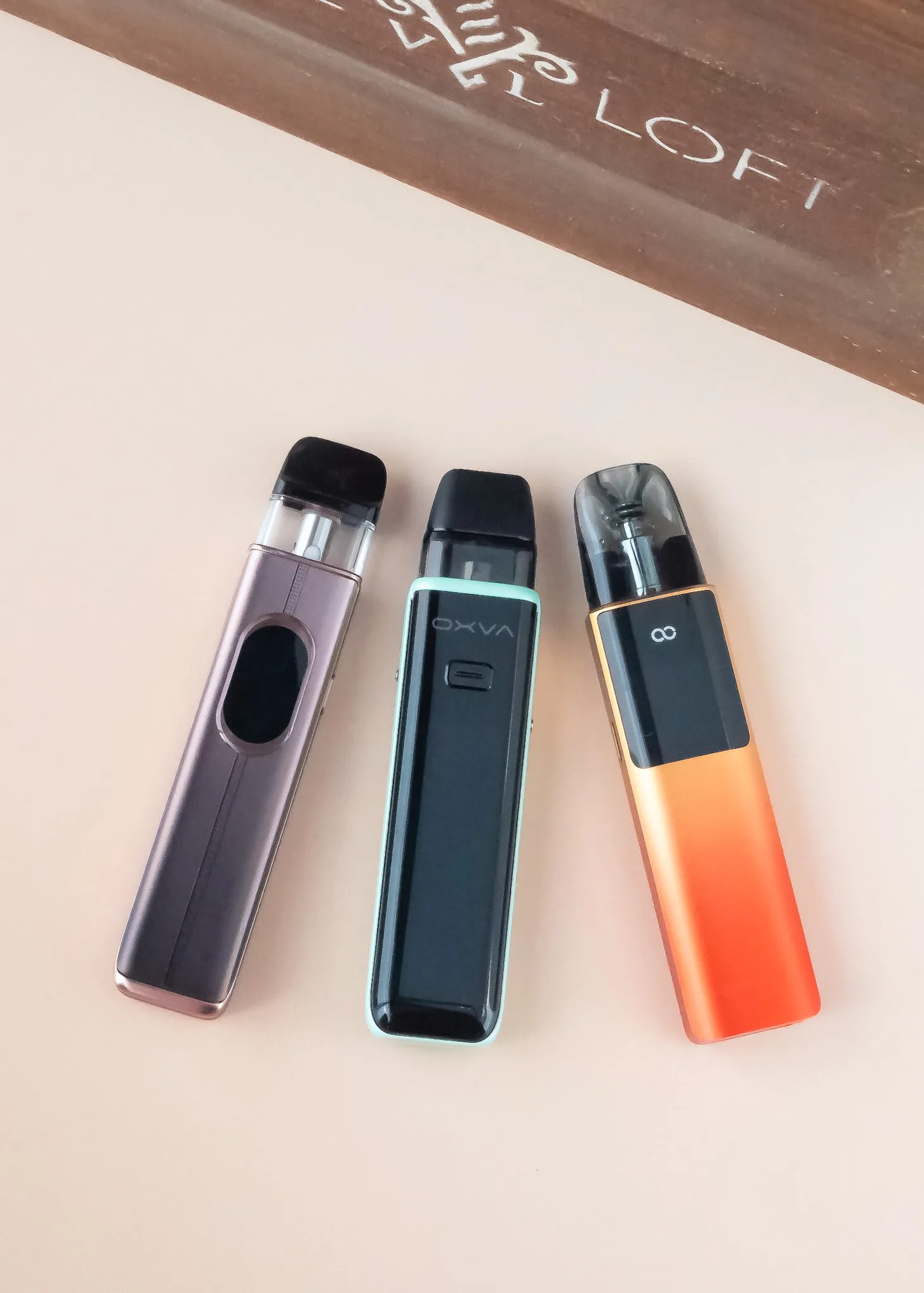 Three smart pod vapes in various colors displayed on a light surface, with a wooden background.