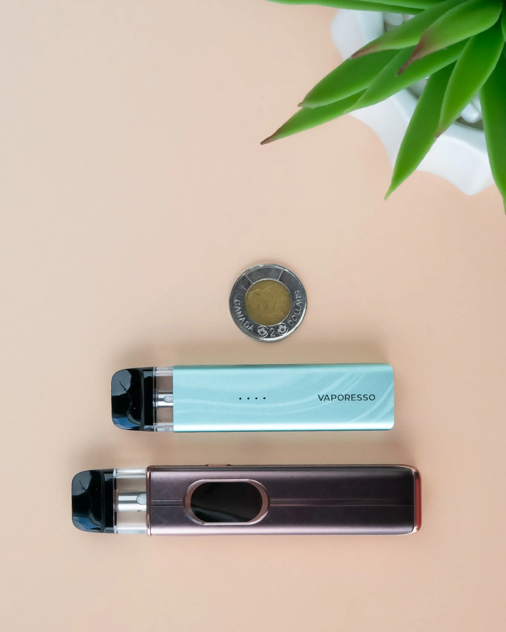 Two pod vapes in mint green and metallic brown, with a coin placed between them on a peach background.